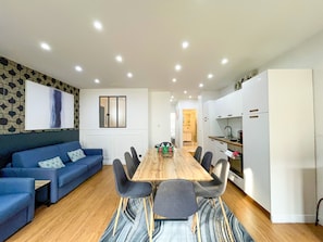 A modern living room with a blue sofa, a wooden dining table with chairs, and a kitchen area with white cabinets and a refrigerator. - UrbanFlat 17 - Pretty 3 Bedroom Flat in Paris Center (París)