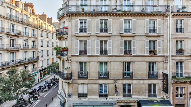 A multi-story building with balconies, windows with shutters, and a street with parked vehicles.
