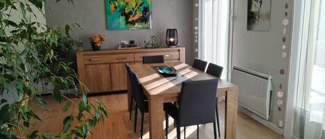 A modern dining area with a wooden table and chairs, a kitchen in the background, and a large window with curtains.