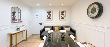 A modern dining area with a long black table, white chairs, a sofa with patterned cushions, and a clock on the wall.