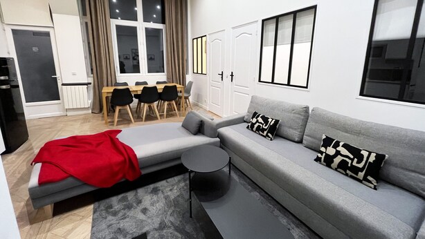A modern living room with a grey sofa, a red blanket, and black and white patterned pillows. There is a dining table with chairs, a window with curtains, and a door with a radiator. - Urban Flat 66 - Pretty 3 bedroom in Paris (París)