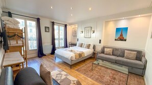 A bedroom with a bed, a grey sofa, a glass coffee table, a wooden dresser, and a window with curtains. - 102 - Urban Opera Picasso (París)
