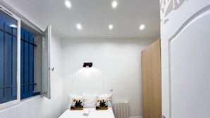 A small, well-lit bedroom with a single bed, a window with blue shutters, and a radiator.