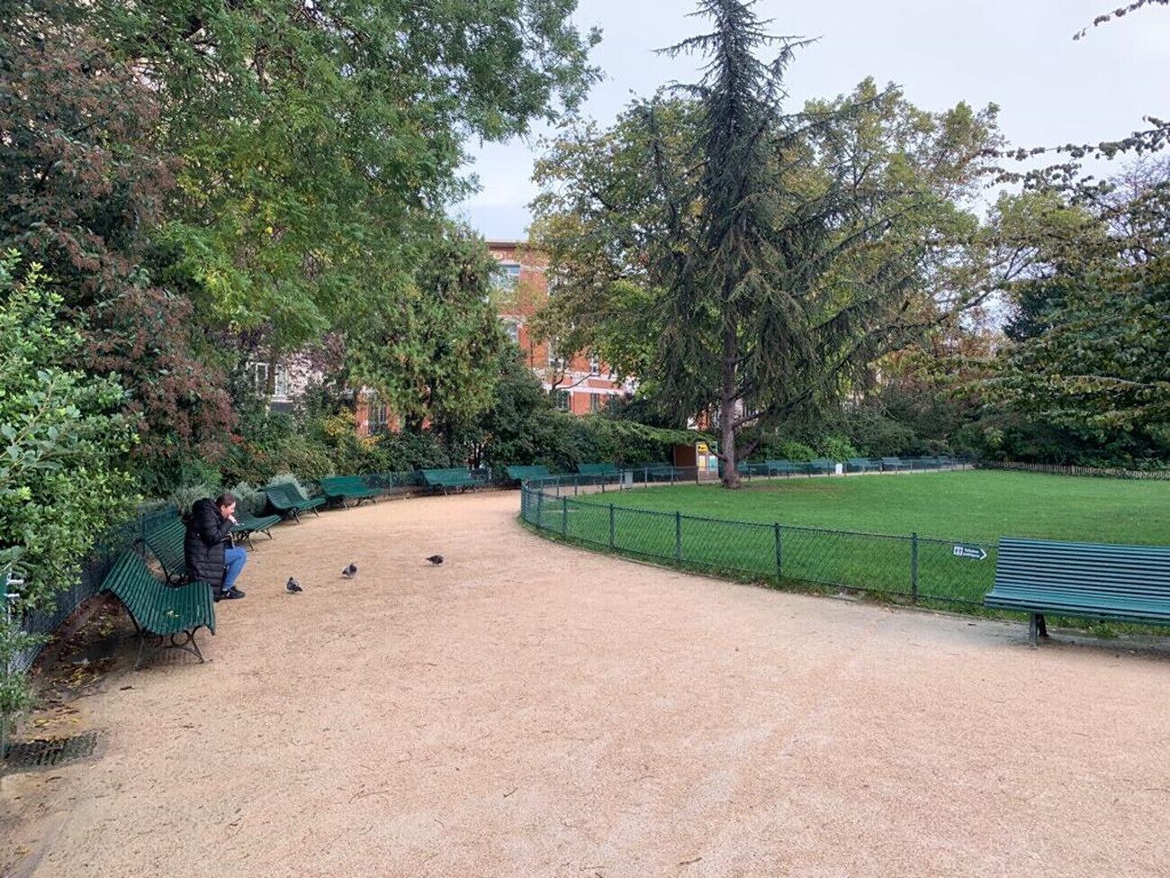 Un parque con bancos verdes, una zona cercada y árboles.