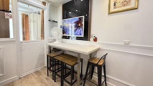 Un área de comedor con una mesa blanca y sillas de madera, un televisor montado en la pared y un cuadro enmarcado sobre la mesa.