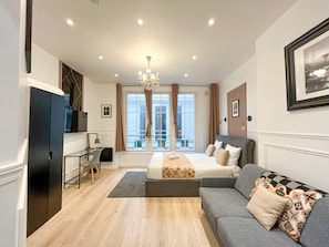 A modern hotel room with a large bed, a sofa, a desk, and a view of the city through large windows. - Urban Flat 55 - 2 Floor Apartment in central Paris (París)