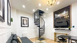A modern living room with a black sofa, a small round table, a black chair, a spiral staircase, and a large TV on the wall.