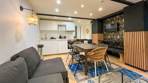 A compact living space with a sofa, dining table, chairs, a kitchen area, and a TV mounted on a wall.