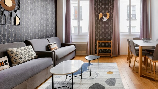 Ein modernes Wohnzimmer mit grauem Sofa, einem kleinen runden Tisch und einem Essbereich mit Holztisch und Stühlen.