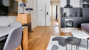 Ein moderner Wohnraum mit Holzboden, einem Flachbildfernseher, einem Essbereich mit Tisch und Stühlen, einer Küche mit Herd und modernen Geräten sowie einem Flur, der in einen weiteren Raum führt.