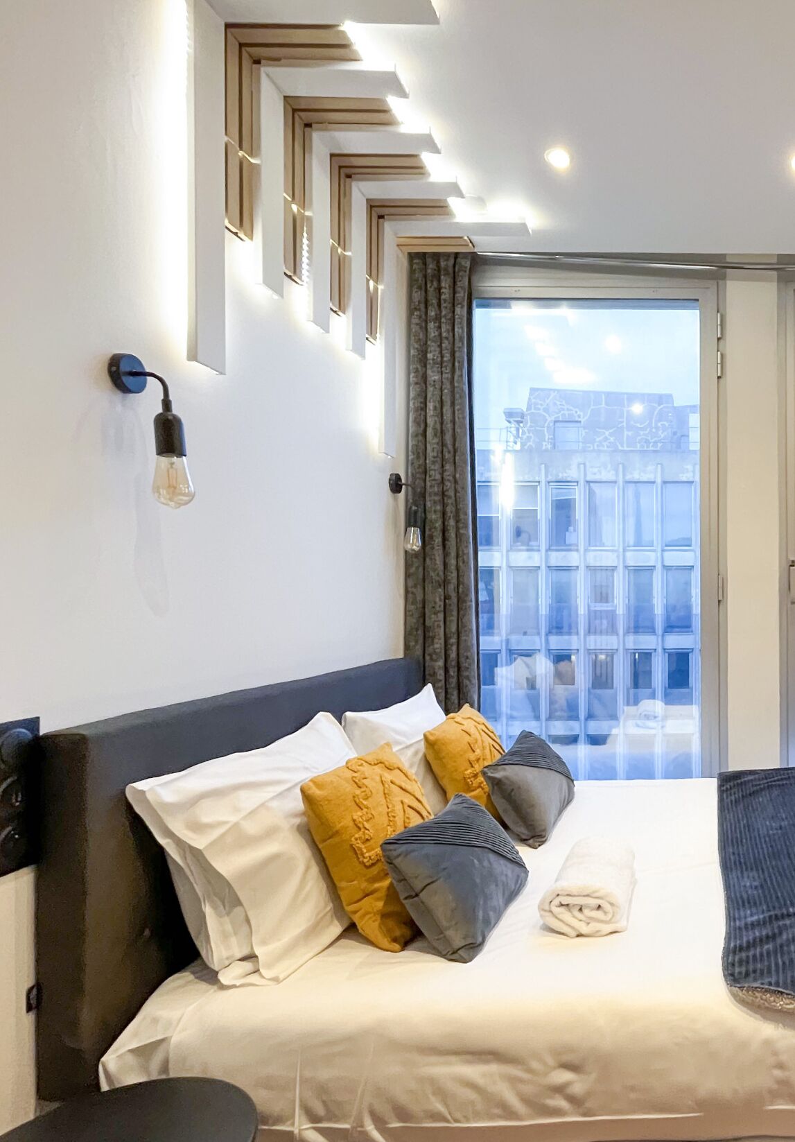 A hotel room with a bed, a wall-mounted light fixture, a window with curtains, and a view of buildings outside.