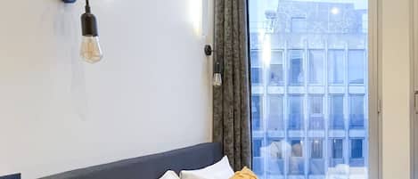 A hotel room with a bed, a wall-mounted light fixture, a window with curtains, and a view of buildings outside.