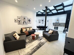 A modern living room with a sofa set, a coffee table, and a dining area with chairs. There is a kitchen visible in the background.