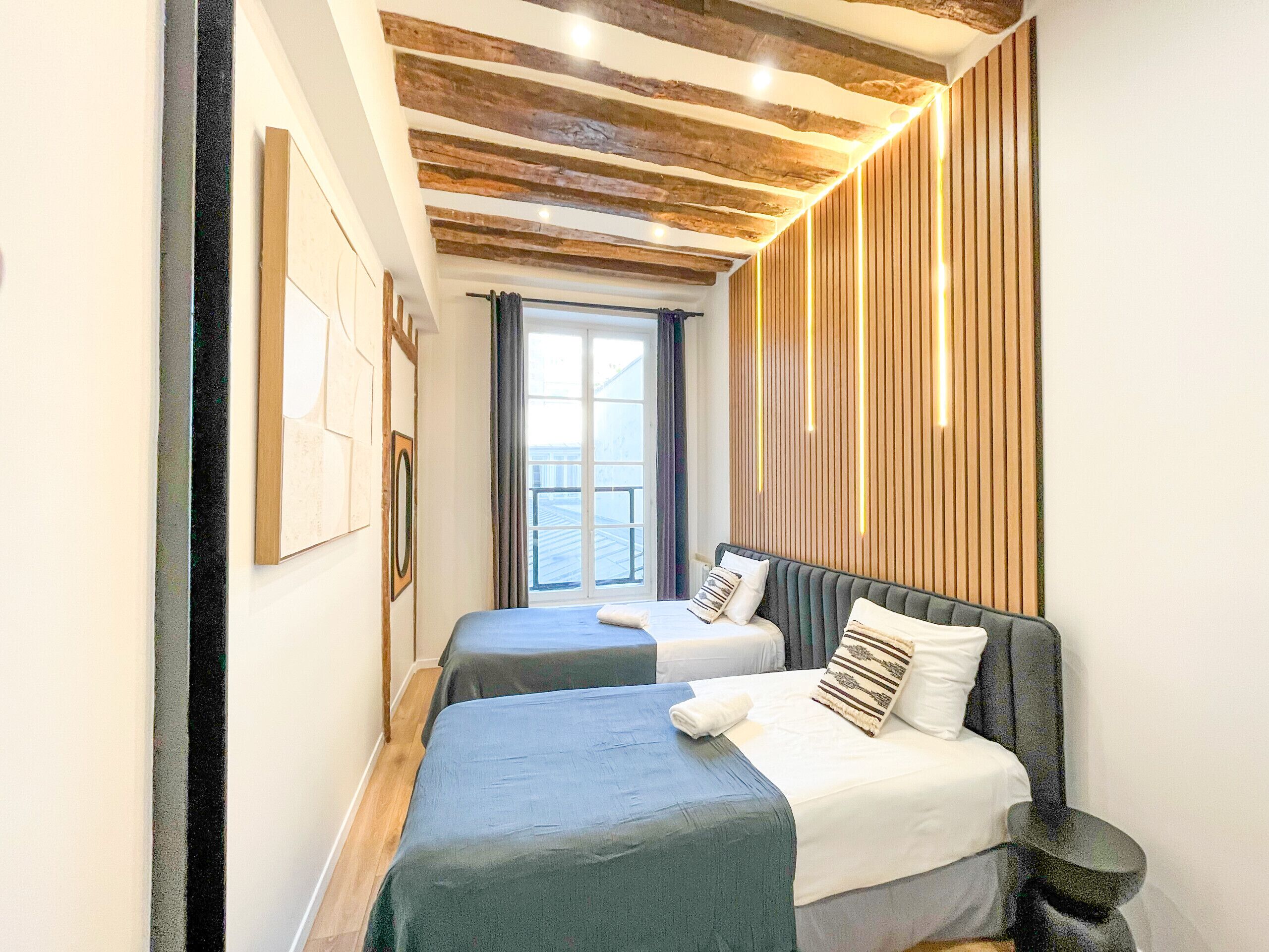 A narrow room with two beds, wooden ceiling beams, a window with curtains, and a wall with vertical wooden slats.