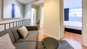 A modern hotel room with a grey sofa, a small black table, and a large painting on the wall. There is a bed with a dark bedspread and a window with curtains.