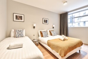 Une chambre d’hôtel avec deux lits, chacun recouvert d’une couette blanche et agrémenté d’oreillers à motifs. Deux tableaux encadrés ornent le mur, ainsi qu’une petite table ronde sur laquelle repose un magazine et une fenêtre avec des rideaux.