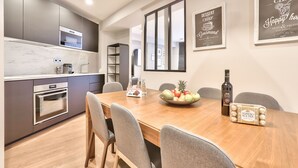 Une cuisine moderne avec une table à manger en bois, des chaises et un panier de fruits. Des tableaux encadrés ornent le mur et une bouteille de vin est posée sur la table.