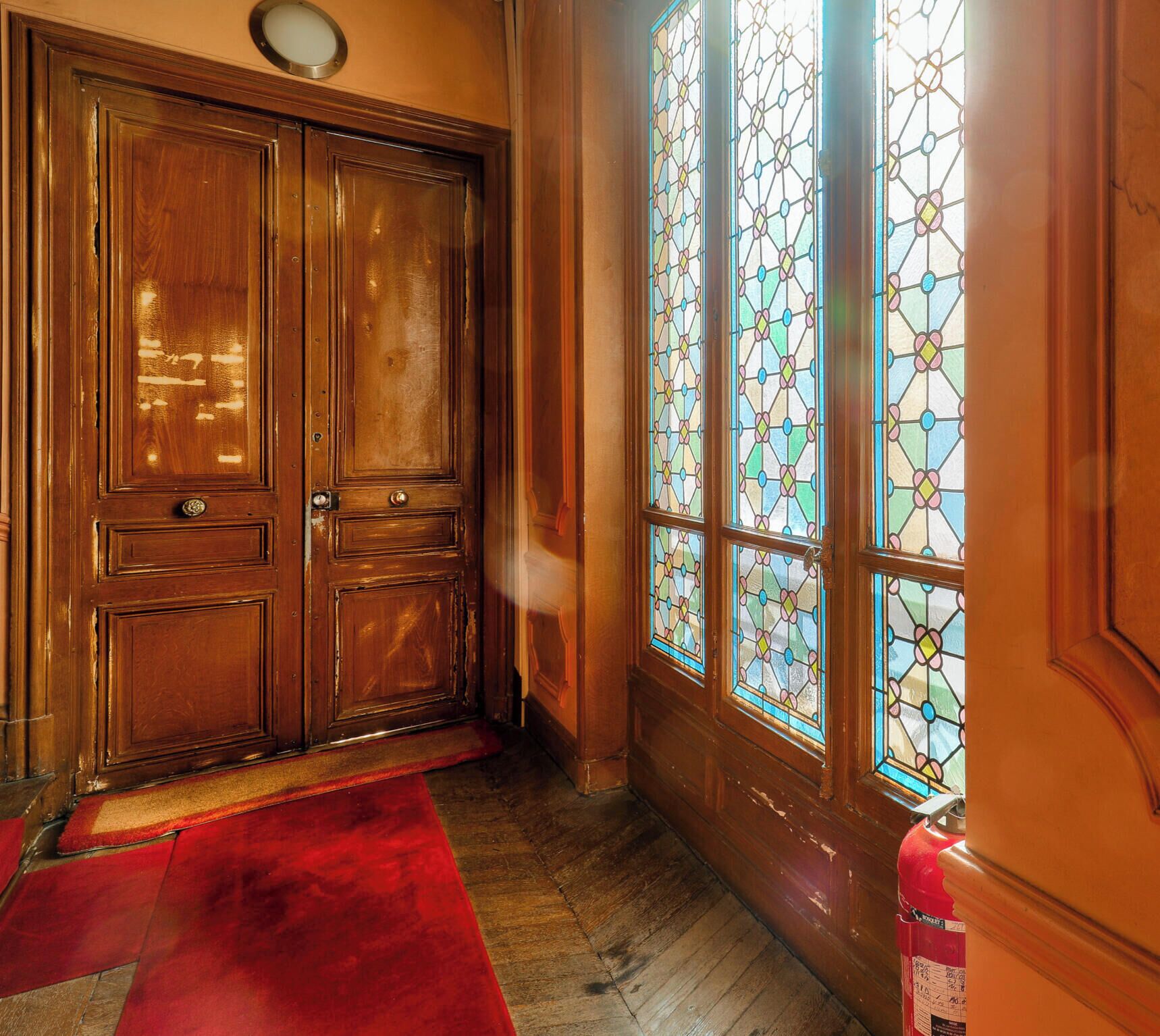 A wooden double door with a red carpet, a stained glass window with a geometric pattern, and a fire extinguisher on the wall.