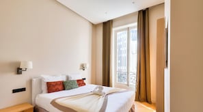 Ein Hotelzimmer mit einem Bett, zwei Wandlampen, einem fensterbelaubten Fenster und Blick auf Gebäude.