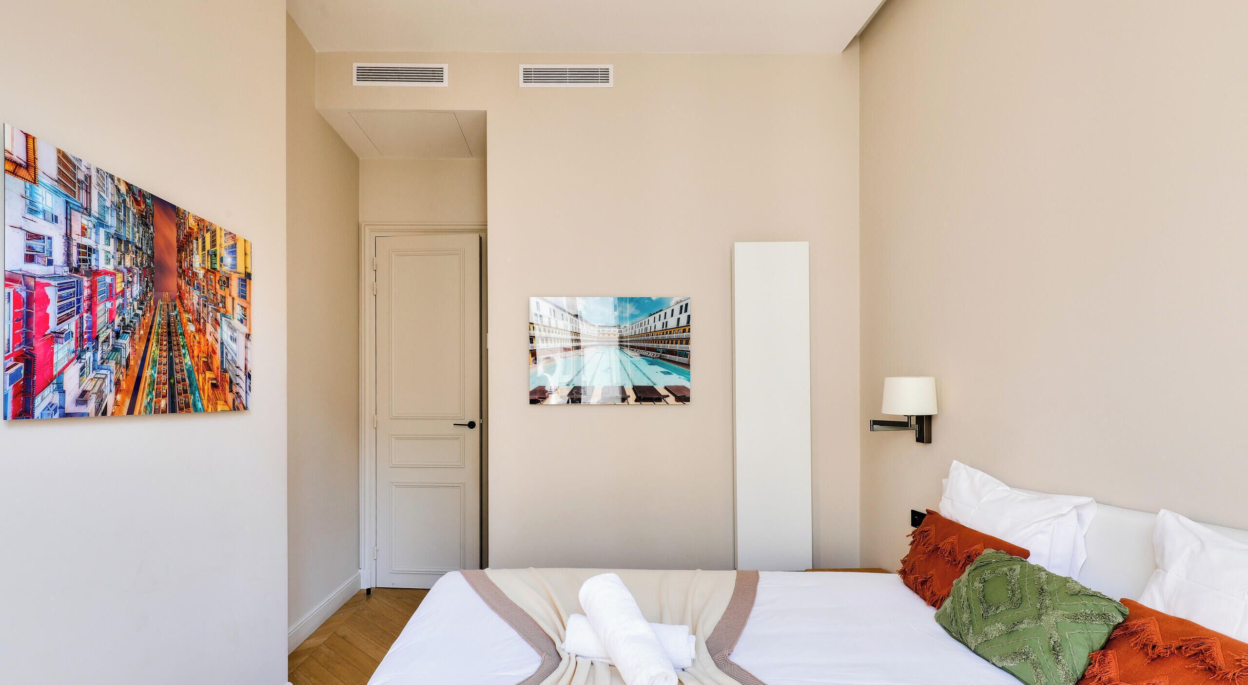 A hotel room with a bed, a painting of a cityscape, a door, and a wall-mounted lamp.