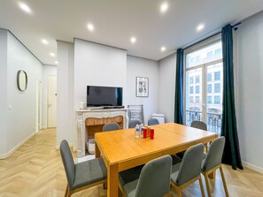 Quarto moderno com uma mesa de jantar de madeira, quatro cadeiras, uma televisão sobre um suporte, lareira e janela com cortinas.