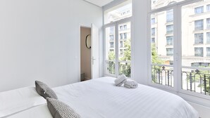 A bedroom with a large window, a bed, and a door leading to another room.