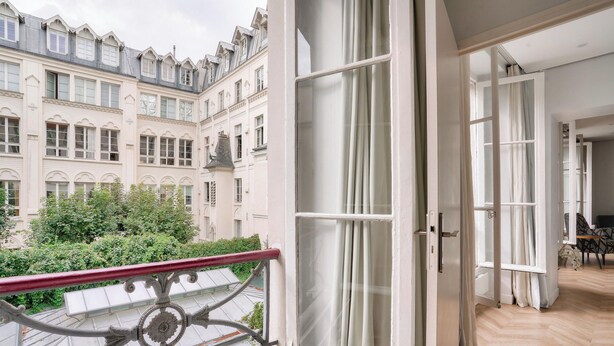 A balcony with a wrought iron railing, a view of a historic building with multiple windows, and a glimpse of an interior space with a chair and a table. - Urban Flat 127 -  4 Bedroom Apartment Paris Montorgueil (París)