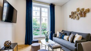 A modern living room with a sofa, ottomans, a coffee table, a flat-screen TV, and a large window with curtains. - UrbanFlat 92 - Prestige Apartment Paris Center (París)