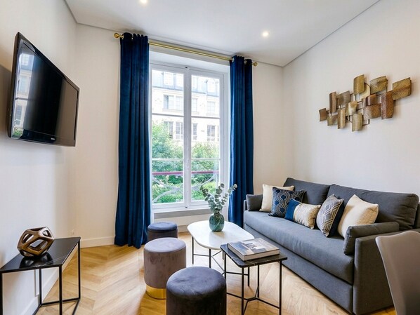 A modern living room with a sofa, ottomans, a coffee table, a flat-screen TV, and a large window with curtains.