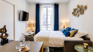 A hotel room with a bed, a TV, a dining table with a plate of macarons, a small table with a candle, and a view of the outside. - UrbanFlat 92 - Prestige Apartment Paris Center (París)