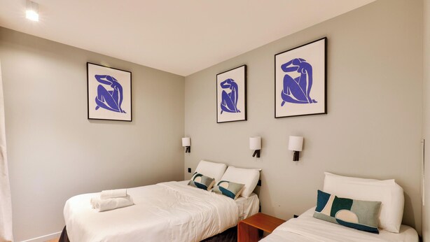 Une chambre d’hôtel avec deux lits, chacun agrémenté de draps blancs et d’oreillers décoratifs. Trois œuvres d’art encadrées ornent le mur, ainsi que deux lampes fixées au mur.