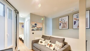 Un salon moderne avec un canapé gris, des œuvres d’art accrochées aux murs et un couloir menant à une chambre à coucher.