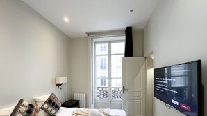 A hotel room with a bed, a TV mounted on the wall, a window with a view of buildings, and a door. - 64 - Urban Luxury Flat near Champs-Elysées (París)