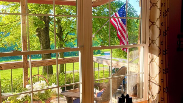 Ein Zimmer mit großem Fenster und Blick auf einen Garten, einer Holzbank und einem Tisch mit einer Blumenkorb.