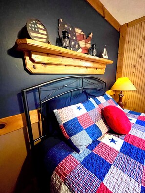 Une chambre à coucher avec un lit recouvert d’une courtepointe ornée d’un motif étoilé, un oreiller rouge et une étagère en bois au-dessus du lit, sur laquelle sont disposés divers objets.