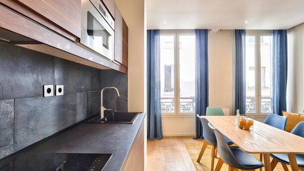 A modern kitchen with dark countertops, a built-in oven, and a sink. There is a dining area with a wooden table and blue chairs. - 71 - Urban Amazing Apartment in Le Marais (París)