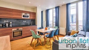 A modern kitchen with wooden cabinets and stainless steel appliances, a dining area with a wooden table and blue chairs, and a balcony with blue curtains. - 71 - Urban Amazing Apartment in Le Marais (París)