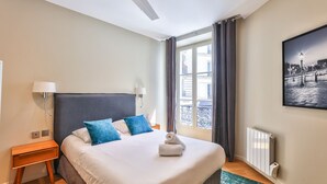 A neatly made bed with blue pillows, a bedside lamp, a radiator, and a window with curtains. - 71 - Urban Amazing Apartment in Le Marais (París)