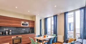A modern kitchen with wooden cabinets and stainless steel appliances, a dining area with a wooden table and blue chairs, and a balcony with blue curtains.