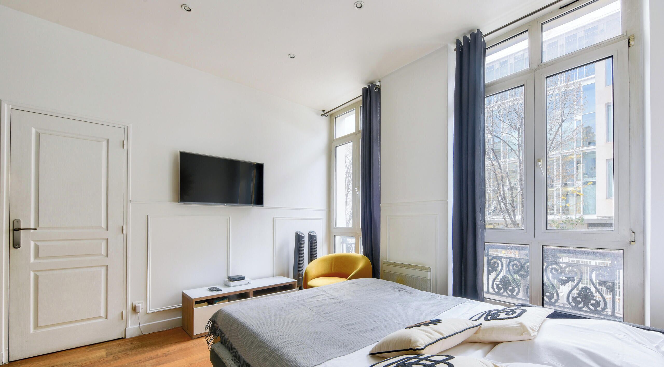 A bedroom with a bed, a desk, a chair, a TV, and large windows.