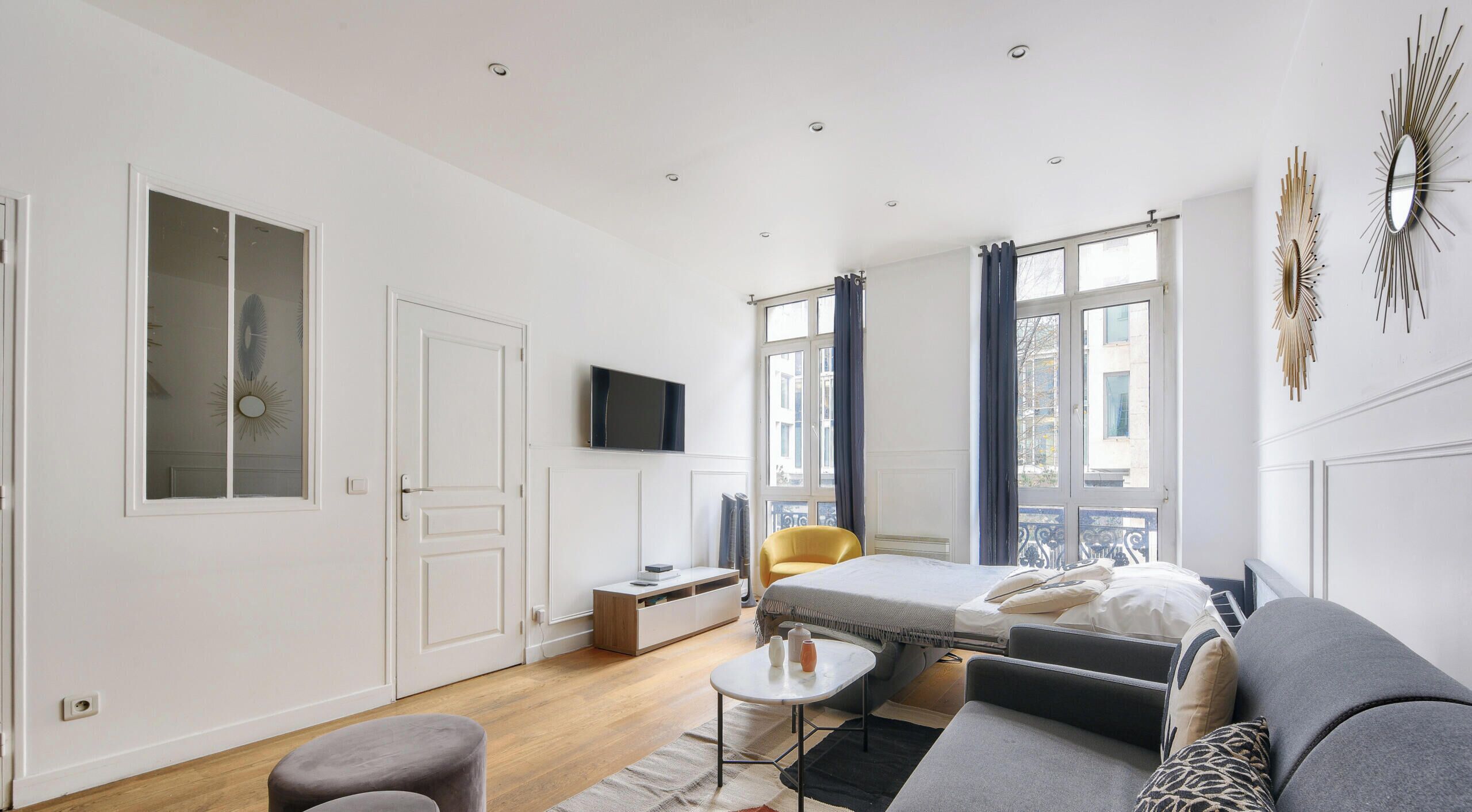 Une chambre d’hôtel moderne, équipée d’un lit, d’un canapé, d’un fauteuil et d’une petite table. On y trouve également un téléviseur fixé au mur, une fenêtre avec des rideaux et une porte dotée d’un miroir.