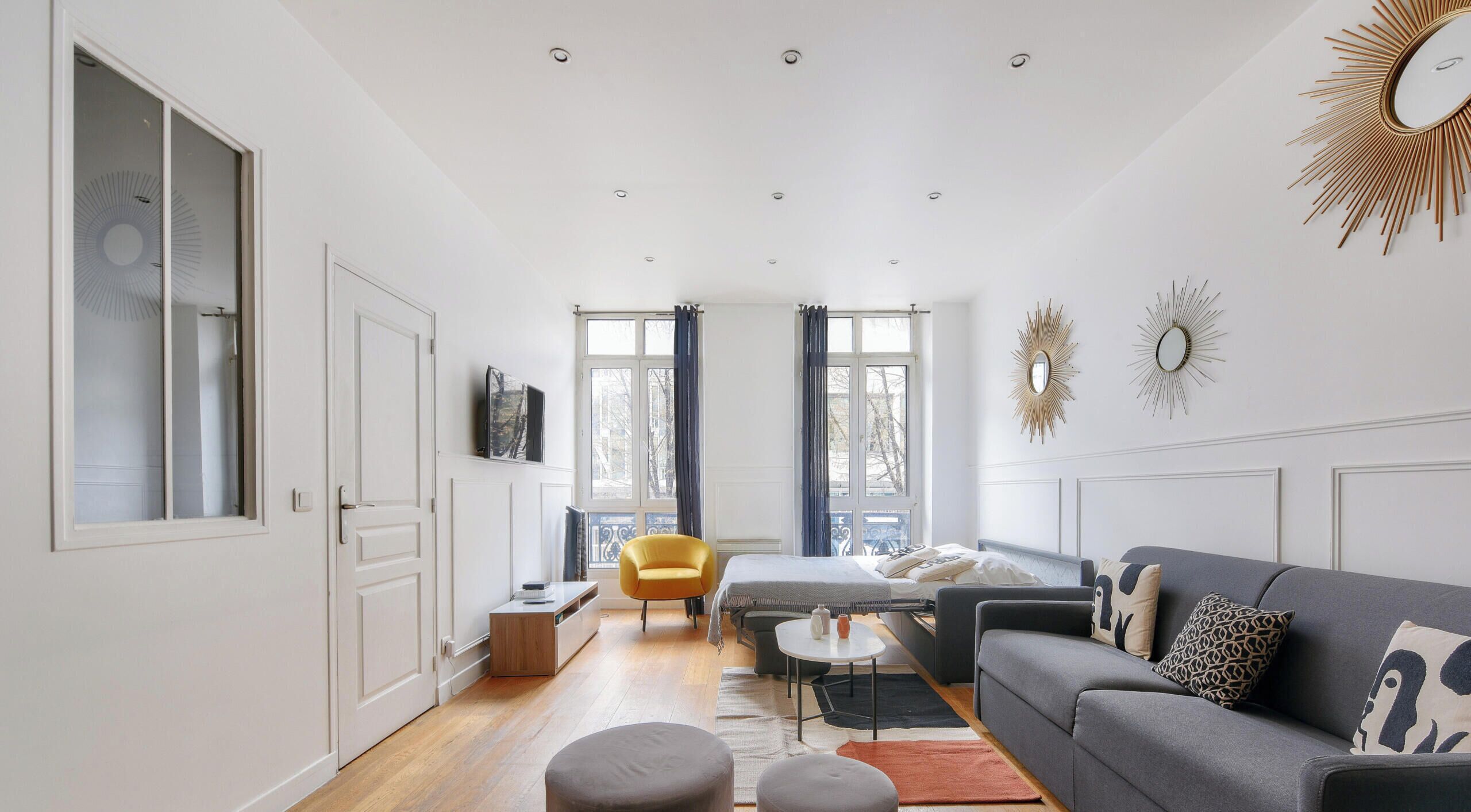 A modern living room with a sofa, armchairs, and a coffee table. There are large windows with curtains, a television on the wall, and a decorative mirror.