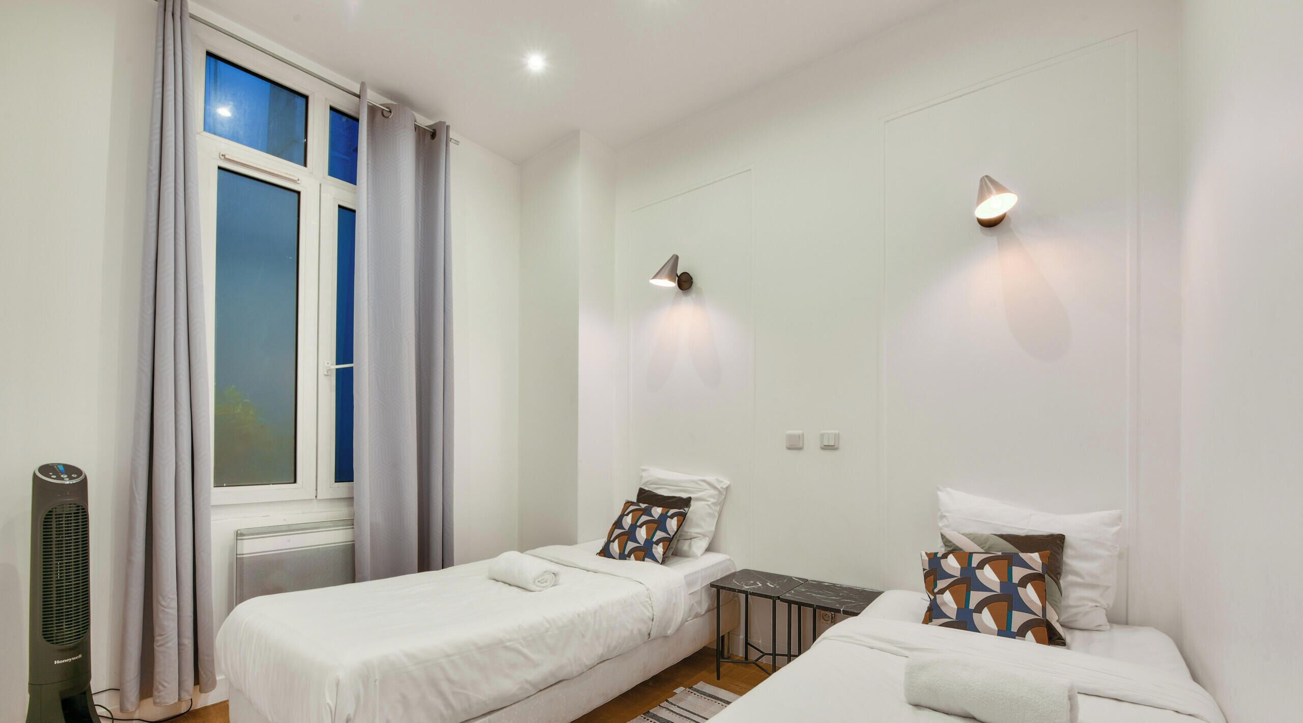 A bedroom with two single beds, a small table, a heater, and a window with curtains.