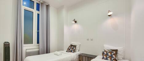 A bedroom with two single beds, a small table, a heater, and a window with curtains.