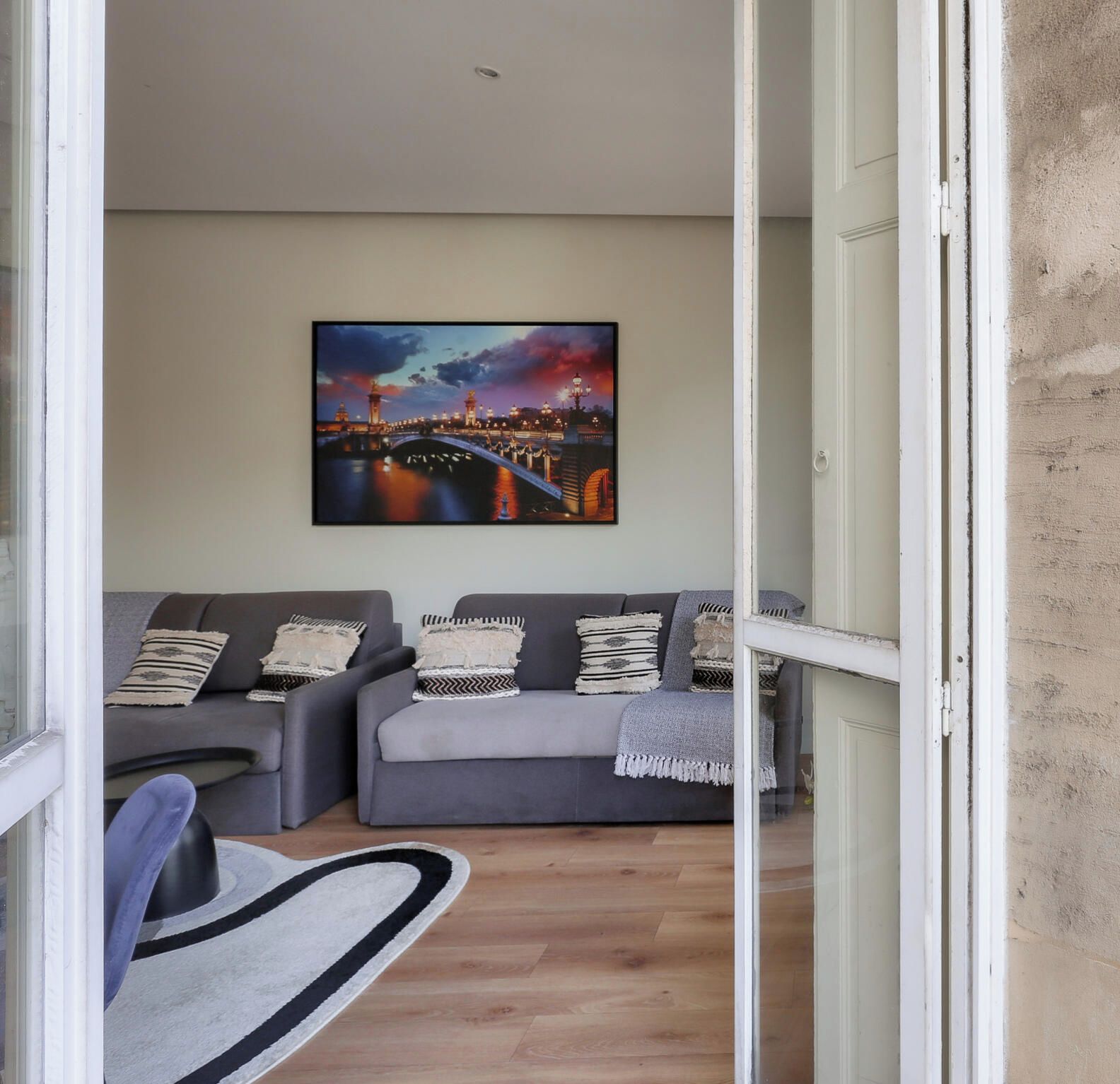A modern living room with a grey sofa set, a large cityscape painting on the wall, and a wooden floor.