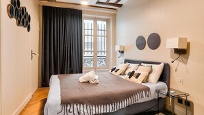 A bedroom with a bed, a window with curtains, a wall-mounted lamp, and a small table.