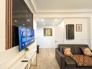 A living room with a flat-screen TV mounted on the wall, a sofa with patterned cushions, a small table with a plant, and a door leading to another room.