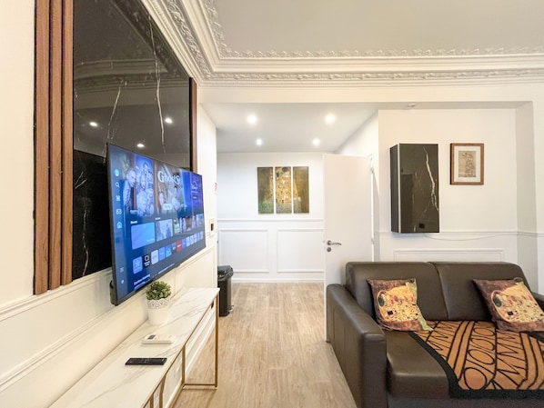 A living room with a flat-screen TV mounted on the wall, a sofa with patterned cushions, a small table with a plant, and a door leading to another room.
