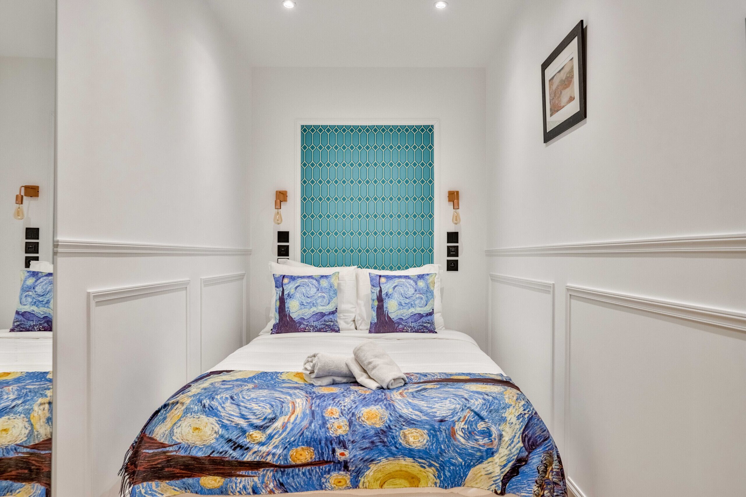Een hotelkamer met een bed voorzien van een sprei met blauw en geel patroon, een geometrisch wandpaneel in teal en twee wandlampen.