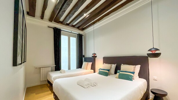 A hotel room with two beds, wooden flooring, a modern ceiling, and a window with curtains. - Urban Flat 69 - Beautiful Parisian Apartment in Center of Paris (París)
